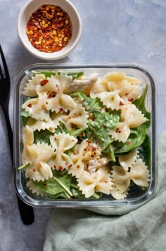 a glass container filled with pasta salad next to a bowl of seasoning and spoon