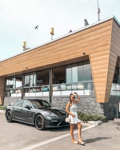 a woman standing next to a black car in front of a building with a wooden roof