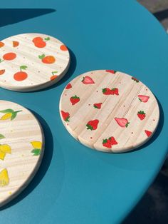 three wooden coasters sitting on top of a blue table next to each other with strawberries and lemons painted on them