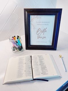 an open bible next to a framed book with writing utensils and pens on it