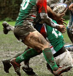 a group of men playing a game of rugby on muddy grass with trees in the background