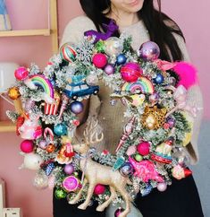 a woman is holding a christmas wreath with ornaments and decorations on it, while standing in front of a pink wall