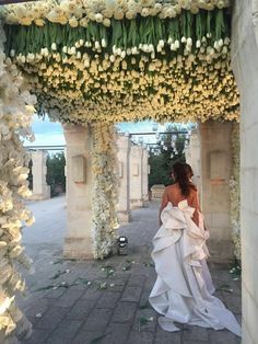 a woman in a white dress is walking under an archway with flowers on the sides