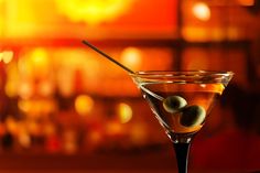 two olives in a martini glass on a bar with blurry lights behind it