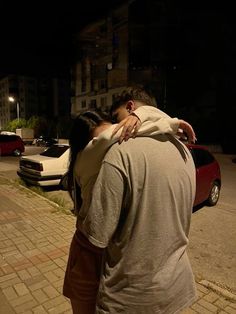 a man and woman hug on the street at night time as cars are parked in the background