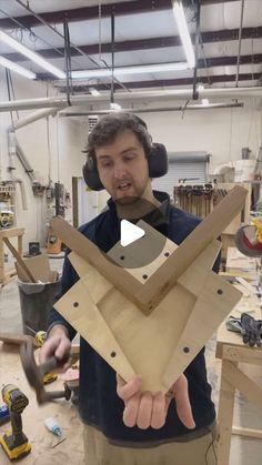 a man holding up a piece of wood with headphones on his ears in a workshop