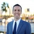 a man in a suit and tie standing by the water with palm trees behind him