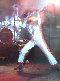 a man in white pants and red shirt playing guitar on stage with other instruments behind him