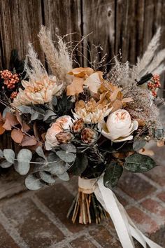 a bridal bouquet with dried flowers and greenery on a brick floor in front of a wooden fence