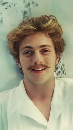 a man with long hair and a moustache smiles at the camera while wearing a white shirt