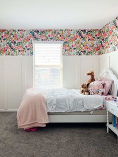 a white bed sitting under a window next to a wall covered in floral wallpaper