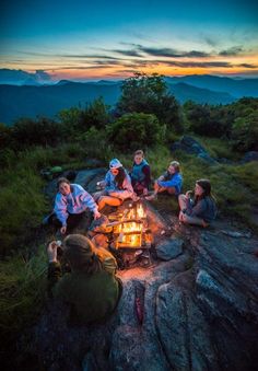 a group of people sitting around a campfire on top of a mountain at sunset