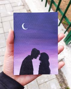 a hand holding up a small card with a painting of two people kissing in front of the moon