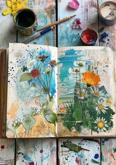 an open book sitting on top of a wooden table covered in paint and paper flowers