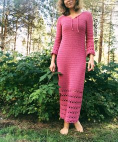 a woman wearing a pink crochet dress standing in front of bushes and trees