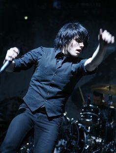 a male in a black shirt is on stage with his arms out and drums behind him