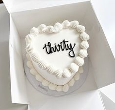 a white heart shaped cake with the word thirty written on it in black lettering, sitting in a box