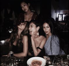 three women sitting at a table with food and drinks in front of them, posing for the camera