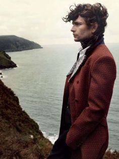 a man standing on top of a cliff next to the ocean with his hands in his pockets