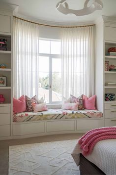 a window seat in the corner of a room with white curtains and pink pillows on it