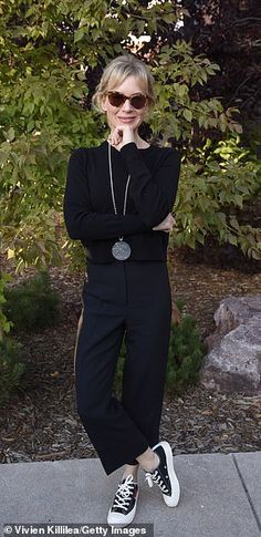 a woman standing on the sidewalk with her arms crossed, wearing sunglasses and a black sweater