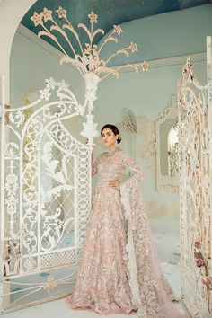 a woman standing in front of a white gate wearing a pink and gold gown with intricate detailing