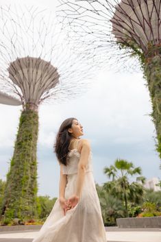 a woman in a white dress standing next to two large trees with plants growing on them