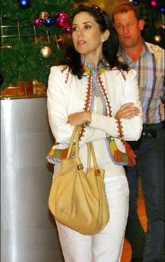 a woman in white pants and jacket carrying a tan purse while standing next to a christmas tree