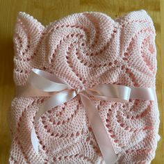 a pink crocheted blanket with white ribbon and bow on it, sitting on top of a wooden table