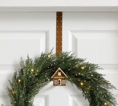 a christmas wreath with a gingerbread house hanging from it's side and lights on the front door