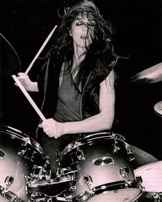 a woman with long hair playing drums in a black and white photo, while wearing a leather vest