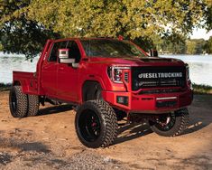 a red truck parked next to a tree in the dirt near some water and trees