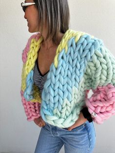 a woman with grey hair wearing a colorful knitted sweater and jeans, standing in front of a white wall