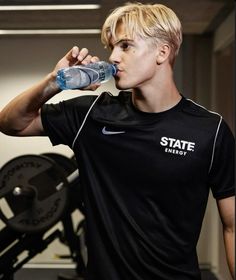 a young man drinking water from a bottle while standing in front of a gym machine