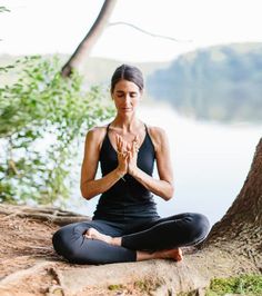 a woman is sitting in the lotus position by a tree with her hands clasped up