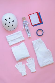 the contents of a doll's white outfit laid out on a pink surface, including gloves and a helmet