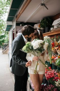 a man and woman are kissing in front of a flower shop while holding bouquets