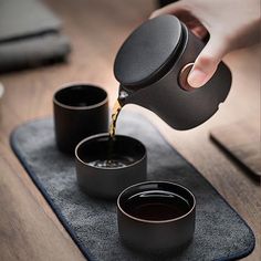 a person pours coffee into three black cups on a mat with a wooden table in the background