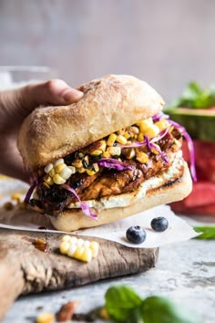 a person holding a sandwich with meat, corn and coleslaw on it next to a cutting board