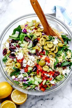 a glass bowl filled with pasta salad next to sliced lemons and olives on a marble surface