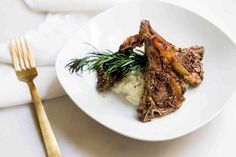 a white plate topped with meat and mashed potatoes next to a gold fork on top of a table