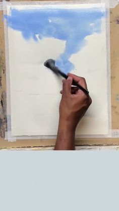 a hand holding a paintbrush and drawing on a piece of paper with blue sky in the background