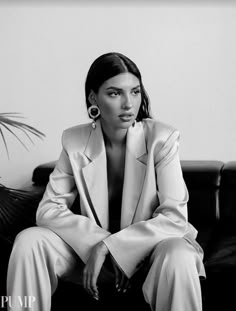 a black and white photo of a woman in a suit sitting on a couch next to a potted plant