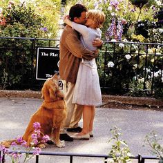 a man and woman kissing while standing next to a dog on a sidewalk with flowers in the background