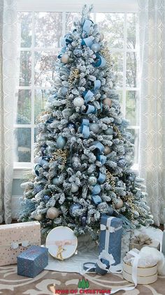 a christmas tree with blue and silver decorations in front of a window, presents on the floor