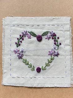 a heart made out of flowers on a piece of white linen with purple thread and green leaves