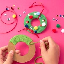 someone making a christmas wreath out of yarn and buttons on a pink surface with other crafting supplies