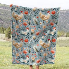 a woman is standing in the grass holding a blanket with roosters and flowers on it