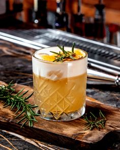 an alcoholic drink with rosemary garnish on the rim and in a glass sitting on a cutting board