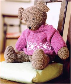 a brown teddy bear wearing a pink sweater sitting on top of a green pillow in front of a wooden chair